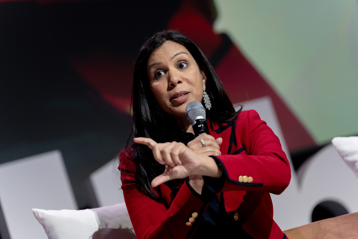 A woman in a red blazer gestures while speaking into a microphone at an event, conveying engagement and enthusiasm.