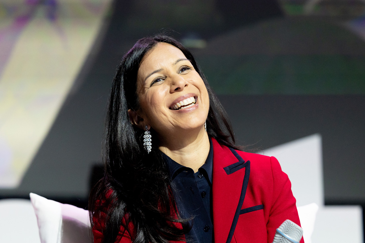 A smiling woman with long dark hair, wearing a red blazer, captures attention with her cheerful expression during an event.