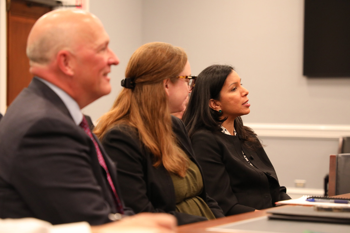 Three individuals seated at a table, engaged in discussion, with a professional setting in the background.