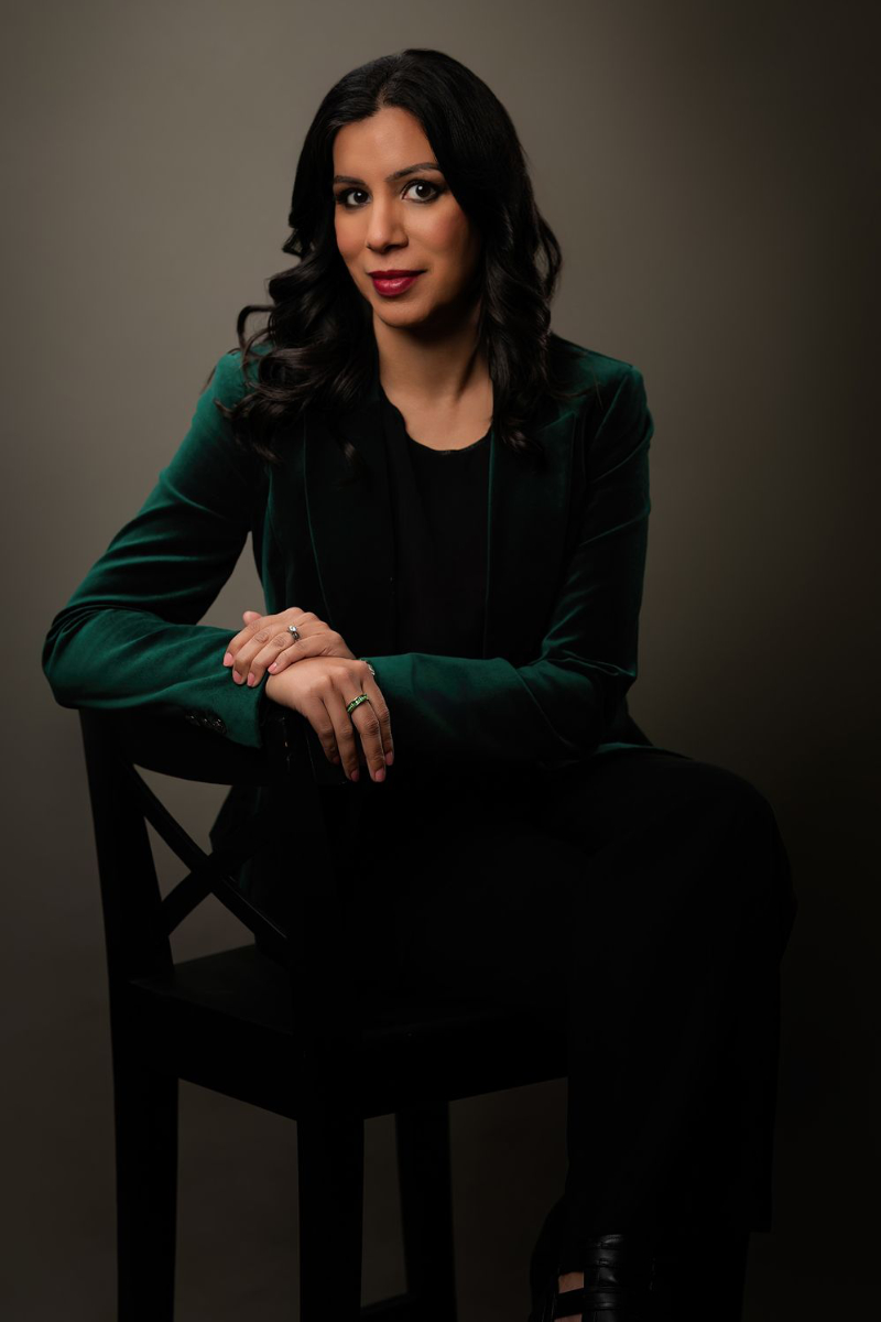 A woman sits elegantly in a dark green blazer, posed thoughtfully on a chair, with soft lighting highlighting her features.