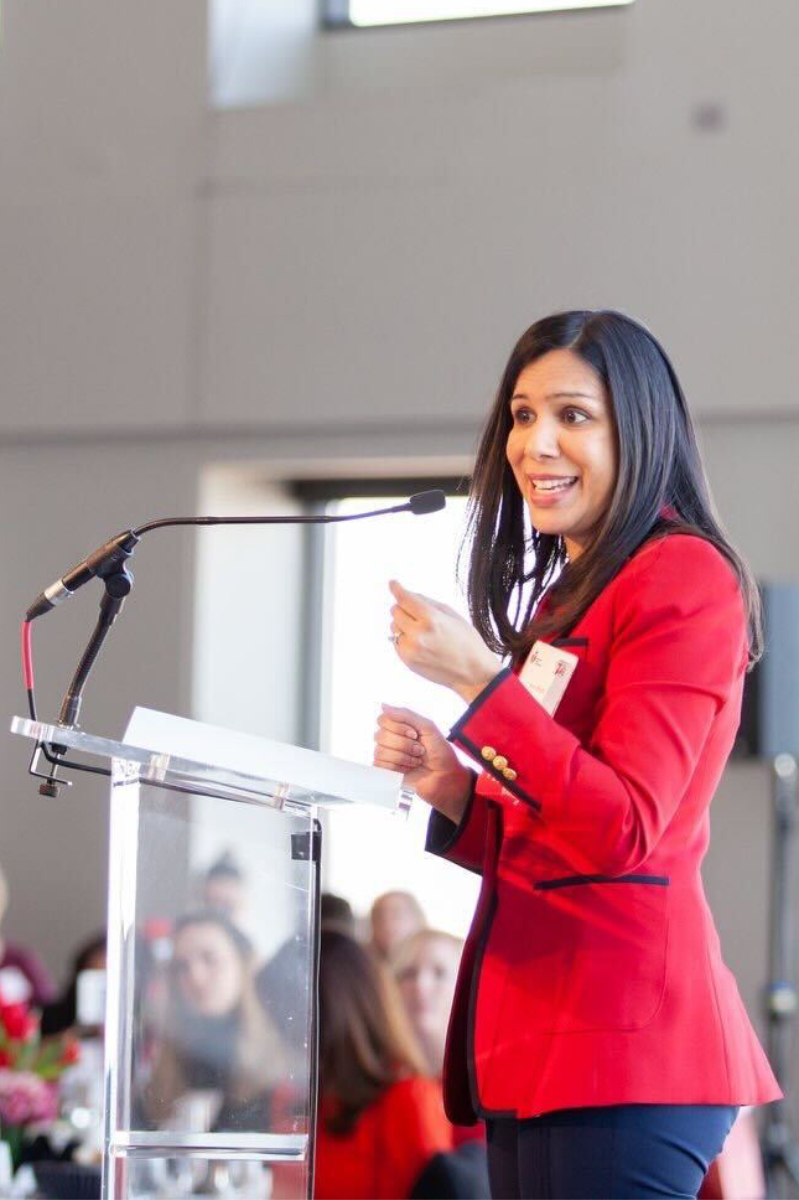 A woman in a red blazer speaks passionately at a podium during a conference, engaging an attentive audience.