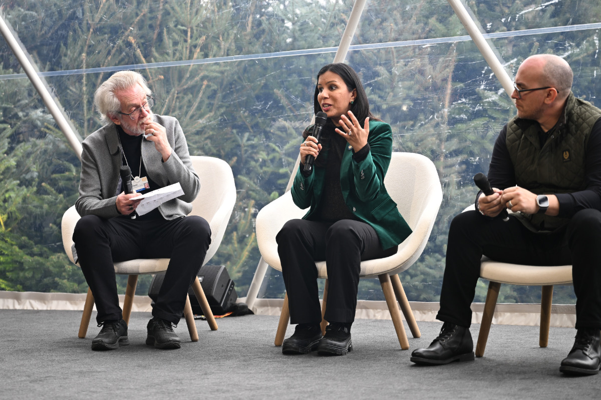 A group discussion featuring three participants seated, engaging in conversation with a forest backdrop visible through the transparent structure.